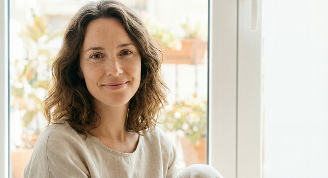 A woman with brown wavy hair and a light-colored shirt smiles gently at the camera while sitting near a window with plants visible in the background.