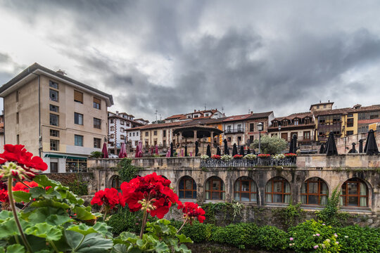 Vista de las terrazas y el templete de Potes en Cantabria, enmarcada por geranios rojos y edificios tradicionales de piedra con arcos bajo un cielo de nubes grises en el coraz&oacute;n de Li&eacute;bana.