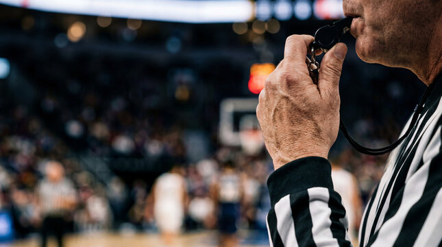 A close-up shot of a basketball referee blowing a whistle during a game, with blurred players and spectators in the background.