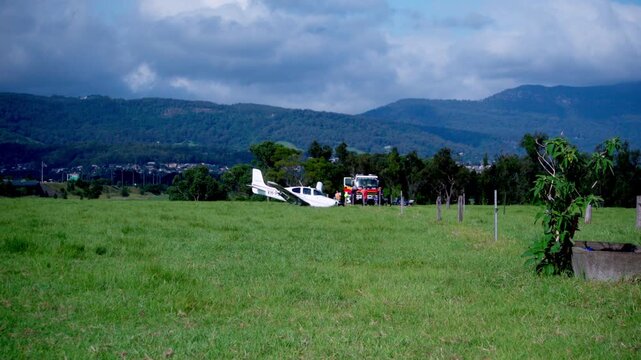 Wide establishing shot of a light aircraft crash site in open rural landscape with emergency services present following crash landing. 