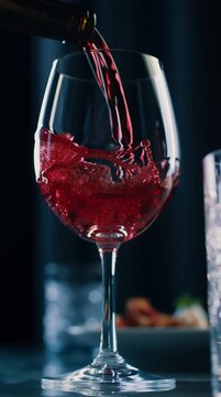 Deep red wine being poured into a clear glass, showcasing the fluid motion and splashes as it fills, with a blurred background of additional glassware and food
