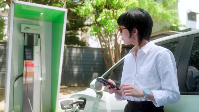A modern connected asian woman checks her smartwatch for charging progress, embodying a tech-savvy and sustainable urban lifestyle beside her EV.