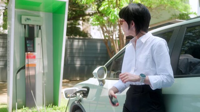 A modern connected asian woman checks her smartwatch for charging progress, embodying a tech-savvy and sustainable urban lifestyle beside her EV.