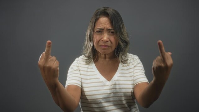 Middle aged woman shows both middle fingers, frowning and glaring at camera in studio with plain gray backdrop and soft directional light; anger defiance.