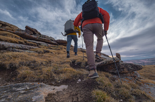 Low angle rear view of two hikers with backpacks and trekking poles climbing up a rocky mountain hill under a cloudy sky, teamwork and outdoor adventure travel concept.