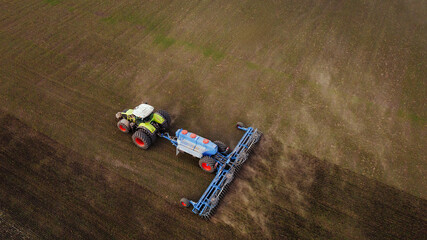 Aerial view of a green and white tractor plowing a brown field with a blue plow attachment, showcasing agricultural machinery in action on farmland © standret