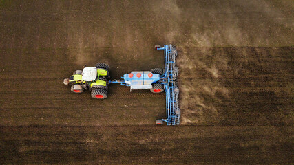 Aerial view of a green and white tractor with orange wheels plowing a brown field using a blue seed drill, showcasing agricultural machinery in action