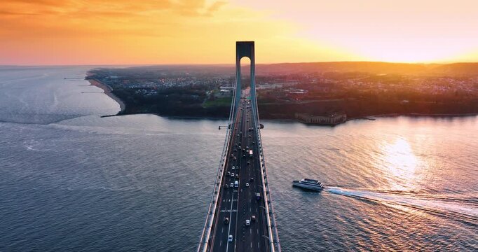 Moving over the multi-lane highways with lively traffic on. A motor boat passes under the Verrazano-Narrows Bridge in New York, USA at sunset.