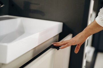 Obraz premium Close-up of a hand reaching for a modern bathroom cabinet drawer with a white sink and faucet in a contemporary interior design setting