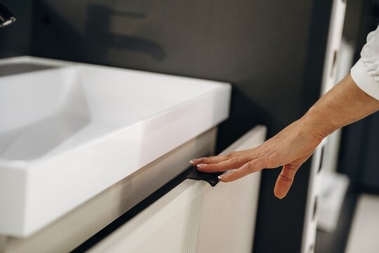 Close-up of a hand reaching for a modern bathroom cabinet drawer with a white sink and faucet in a contemporary interior design setting