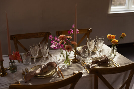 Elegant tablescape with golden cutlery, crystal glasses, and pink flowers on a linen cloth. Warm sunlight illuminates the romantic dinner setting with candles and vintage chairs in a rustic room.