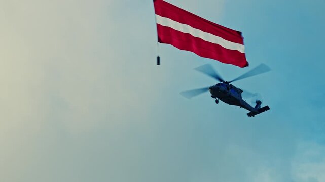 Cinematic low-angle shot of two military helicopters (Sikorsky UH-60 Black Hawk) performing a ceremonial flypast, carrying the red-white-red national flag of Latvia against a dramatic cloudy blue sky.