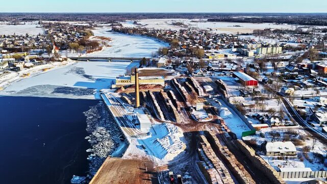 Industrial lumber facility near a town, featuring frozen river and stockpiled logs, Aerial view
