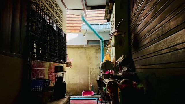 Narrow outdoor kitchen corridor in a residential home with natural light, showing household items, water pipes, and washing area. Everyday domestic lifestyle scene with warm tones and shadow contras