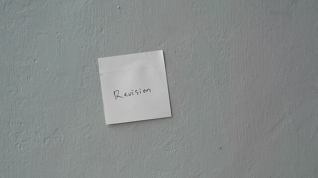 Hand sticking a white square paper note with the word Revision on a textured wall.
