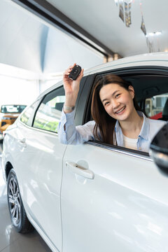 Happy young Asian woman smiling inside a new white car at an auto dealership showroom, proudly showing her new car key. Concept of buying a new vehicle. Vertical Shot
