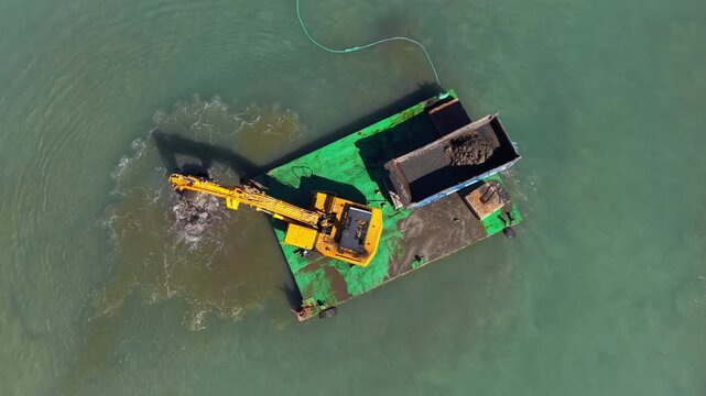 Top-down view of a green dredging barge with a yellow hydraulic excavator removing sediment from shallow coastal waters, loading material into onboard containers