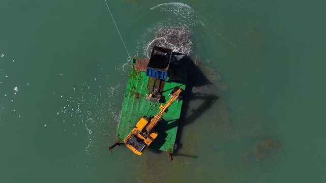 Top-down view of a construction barge with yellow excavator working on land reclamation in turquoise green sea water, pumping sediment to create artificial island