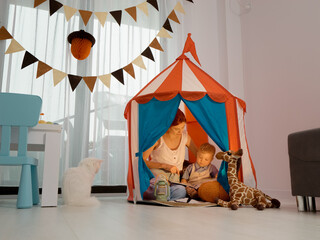 In a colorful tent at home, a mother reads a storybook to her young son while their cat sits beside them, creating a warm and cozy atmosphere. © Binzari