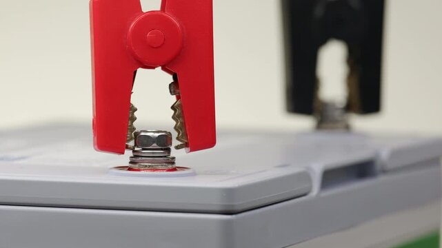 Battery charging process begins as red positive clamp connects to battery terminal. Black negative connector already attached. Metal clamps secure electrical connection for power transfer.