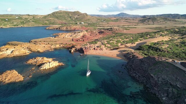 Aerial drone view around a yacht, in the bay of the Cala Pregonda, Menorca Island, Balearic islands, Spain. Red sandy rocks in the background