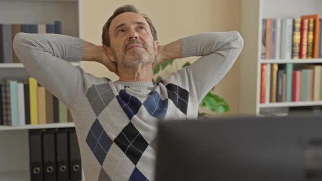 Man reclining with hands behind head at computer in building, eyes closed and leaning back at desk, wearing argyle sweater and beard visible; calm break.