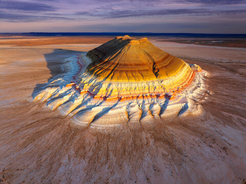Aerial view of the striking multi-colored rock formation casts long shadows over the arid landscape, its layered strata painted in hues of yellow, orange, and white, Aktau, Kazakhstan.