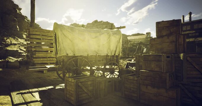 In a rustic frontier environment, a covered wooden wagon stands among stacked crates, surrounded by dusty terrain and dramatic clouds in the afternoon light.