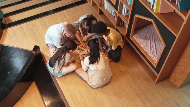 Group of diverse children sitting in circle while reading a book at library. Top view of girl sharing a magazine while pointing at interested topic and talking with multicultural friends. Edification