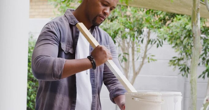 African American man mixing white paint after opening bucket, preparing roller for patio painting