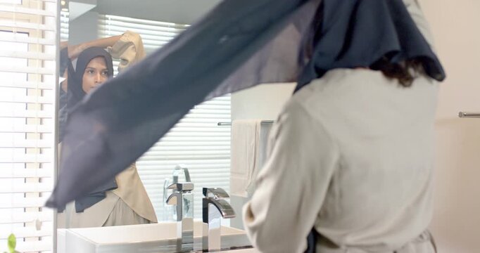 African American woman inspecting mirror, wrapping dark headscarf at bathroom vanity, checking fit