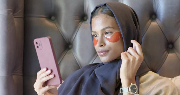 African American woman wearing hijab using phone mirror, applying eye patches on bed for skincare