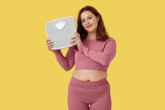 Overweight young happy woman with scales on yellow background. Weight gain concept