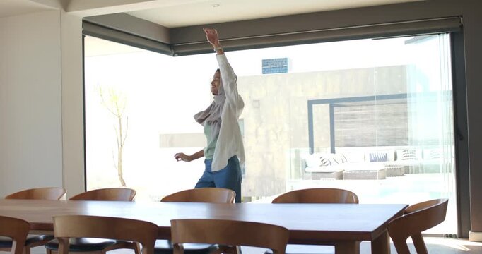 African American woman wearing hijab tapping headphones then dancing behind table near glass door