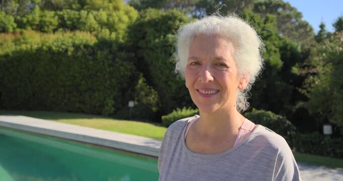 Senior woman standing by pool in gray top with pink strap, camera moving closer, smile widening