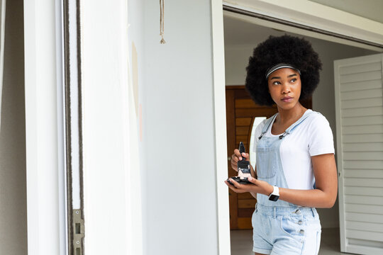 Woman wearing denim short overalls holding paintbrush and paint pot at doorway, copy space