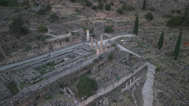 Aerial drone orbit shot around the Temple of Apollo ruins in Delphi, Greece at sunset, showcasing ancient archaeological landmark.