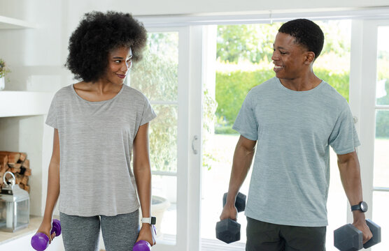 African American couple in gray holding purple and black hex dumbbells, smiling by glass doors