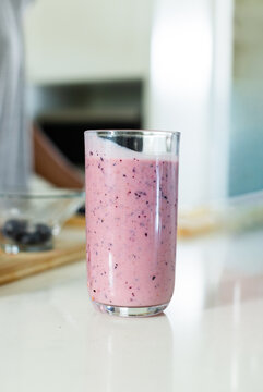 Clear glass sitting on glossy counter holding pink smoothie showing flecks, berry bowl on board