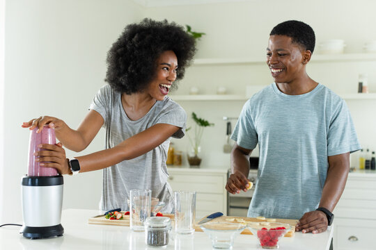 African American couple in kitchen preparing smoothie with blender and slicing fruit with knife