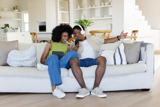African American couple sitting on light-colored sofa in living room, holding tablet and smartphone