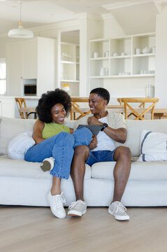 African American couple laughing, sharing tablet on light sofa in living room, woman in green top