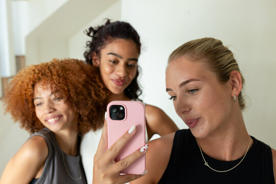 Female friends wearing sleeveless tops taking selfie in apartment with pink phone case, copy space