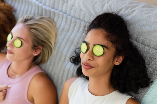 Diverse female friends wearing tank tops lying on ribbed gray blanket at home with cucumber slices