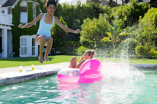 Two female friends, 20s, jumping into home pool in swimsuits splashing pink float and yellow drinks