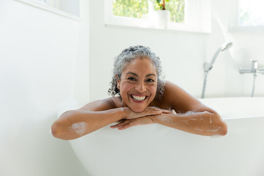 Senior African American woman leaning on freestanding tub rim in bright bathroom, smiling with suds