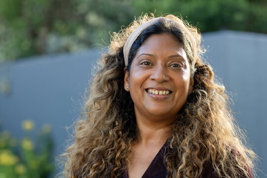 Adult woman smiling and standing in backyard, wearing dark V-neck top with headband, stud earrings