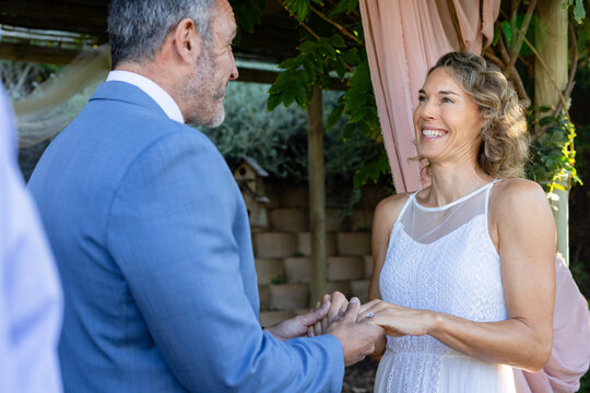 Middle-aged couple exchanging rings under pergola draped with pink fabric, bride wearing lace dress