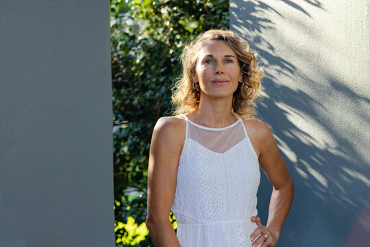 Woman standing in sunlit courtyard in white lace dress, looking as gray walls cast leaf shadows