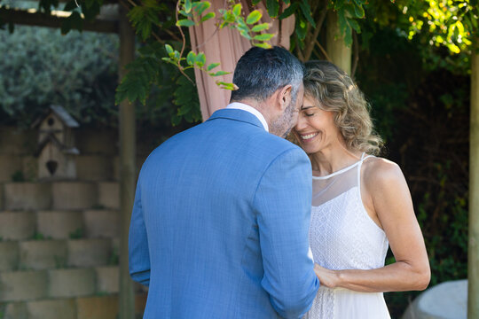 Couple touching under pergola draped with fabric and vines in garden wearing suit and lace dress